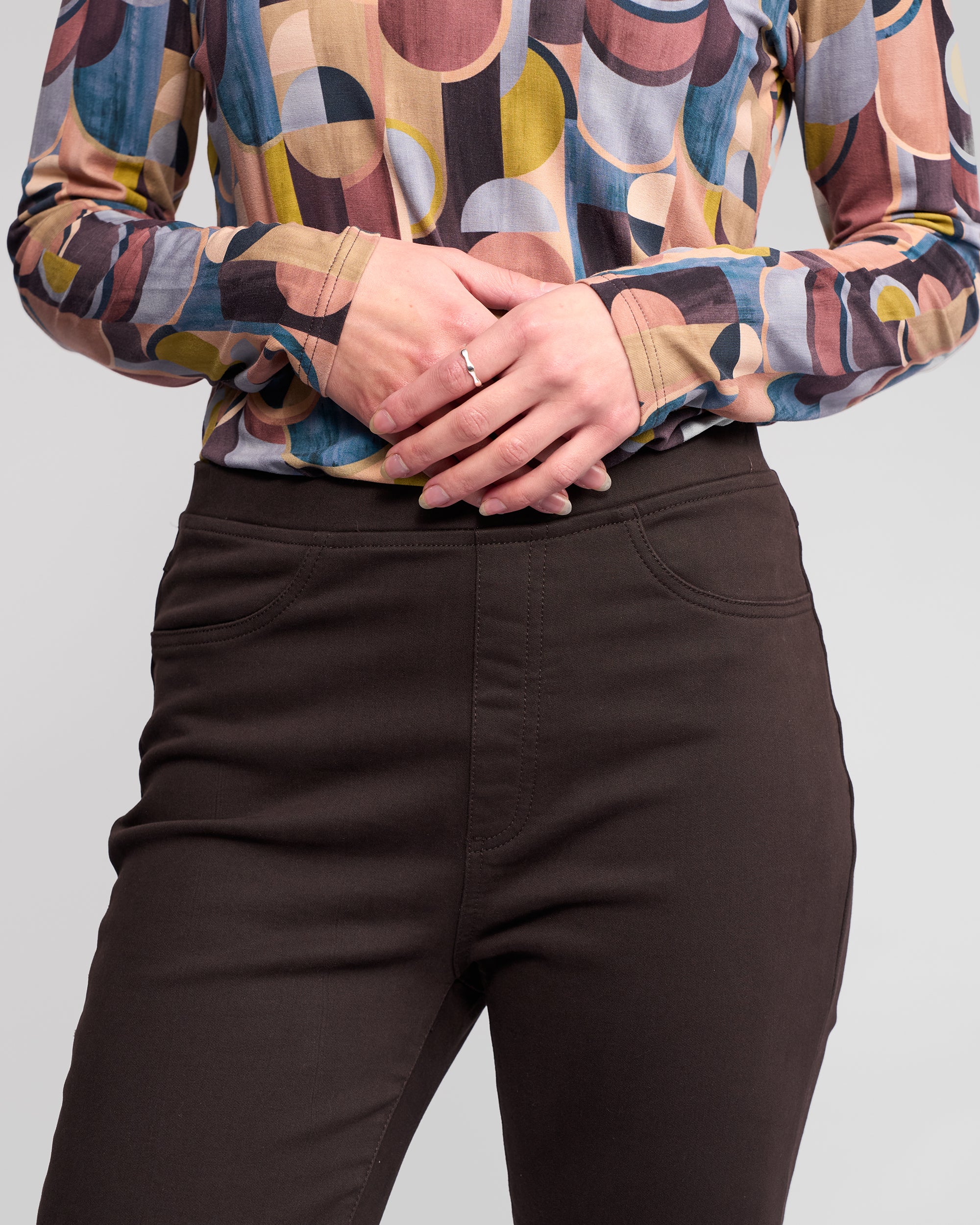 A person wears Vassalli’s Bay Knit Pull On - Chocolate pants with a colorful geometric-patterned long-sleeve top, standing with hands folded at the waist and a thin ring visible. The stretch fabric ensures comfort and style.