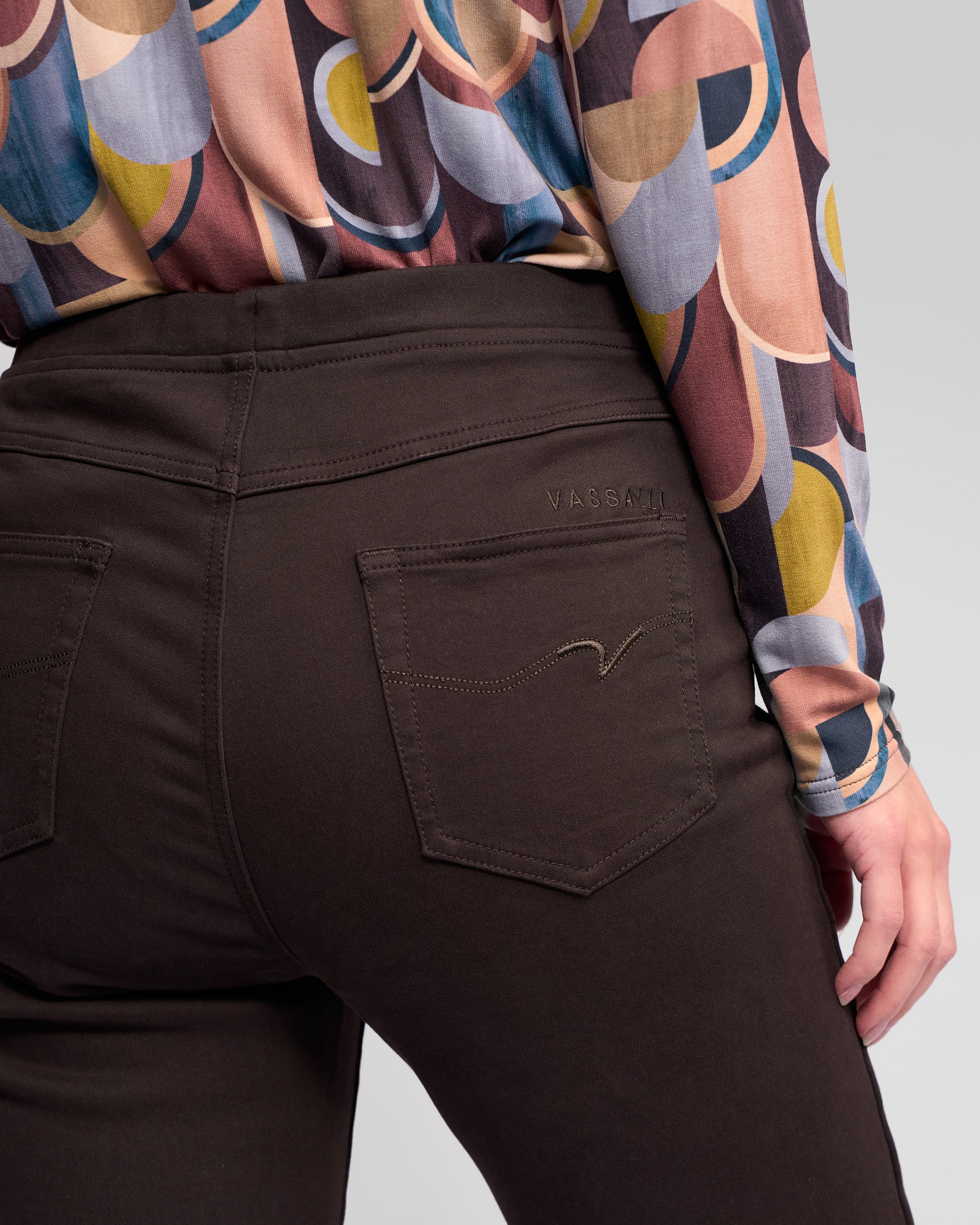 A person wearing Vassalli Bay Knit Pull On pants in chocolate, featuring visible back pockets and brand embroidery above the right pocket. Only their lower back and hand are shown, paired with a colorful geometric-patterned blouse.