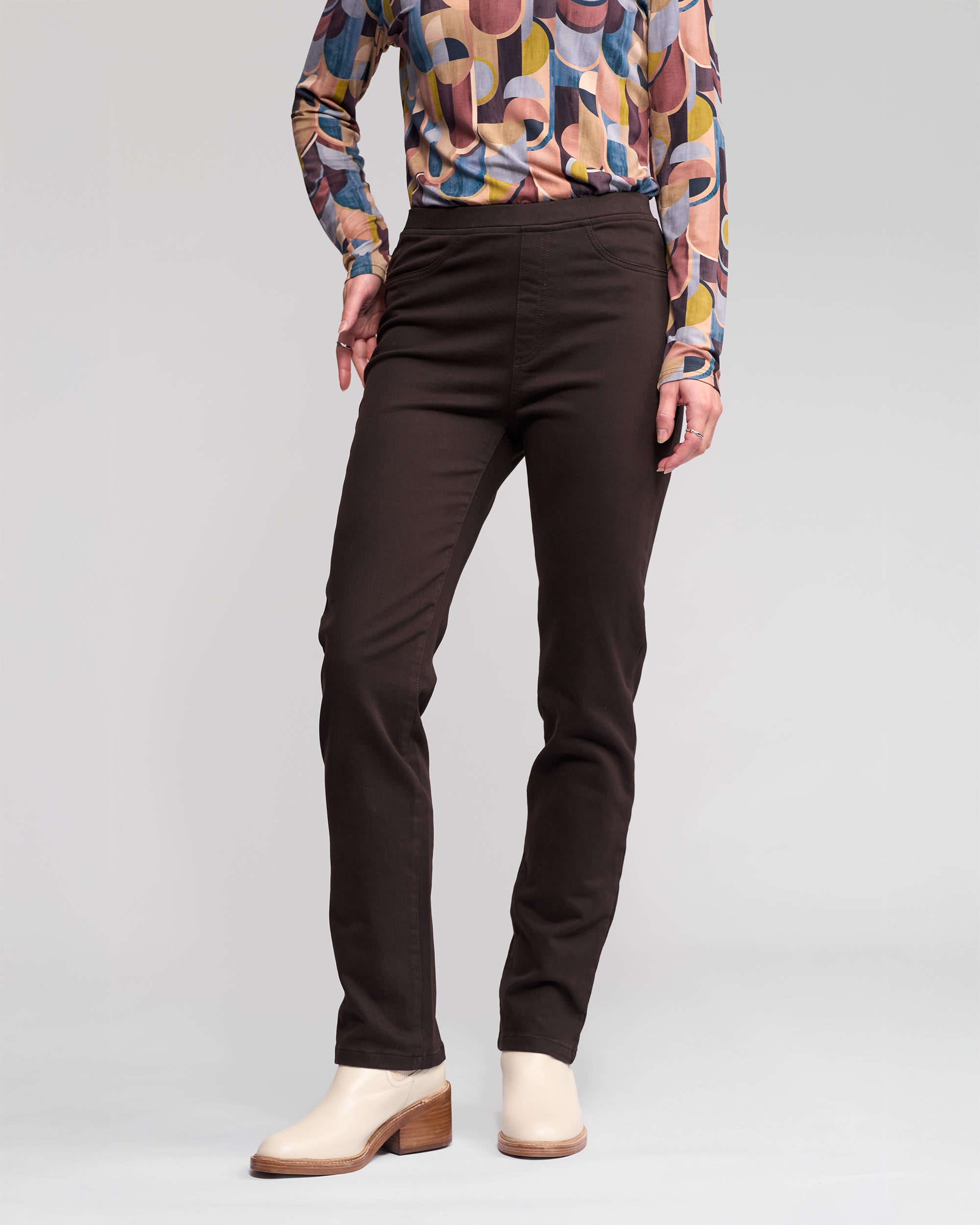 A person in Vassalli Bay Knit Pull On pants in chocolate stands against a plain light background, wearing a colorful geometric long-sleeve top and beige heeled ankle boots. The person's head is not visible.