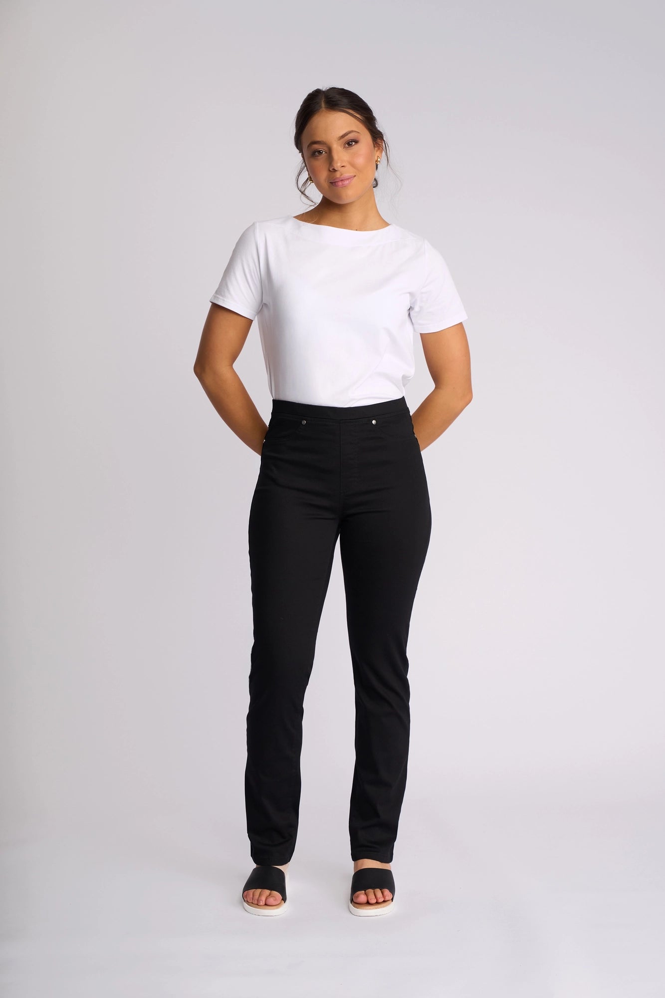 A woman stands against a plain white background, wearing a white short-sleeve shirt and black slim-leg pants—the Bay Pull On by Vassalli—crafted from lightweight stretch fabric, paired with black open-toe sandals, with her hands behind her back.