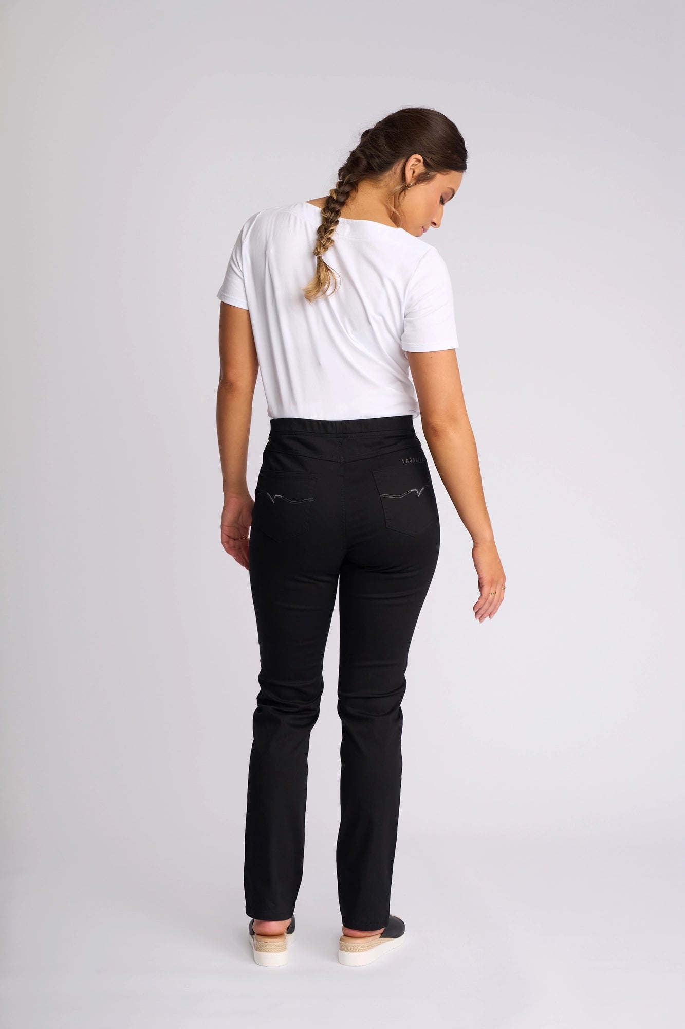A woman with a braid, dressed in a white t-shirt and Vassalli Bay Pull On slim-leg pants in black—crafted from lightweight stretch fabric—and white shoes, stands facing away against a plain white background.