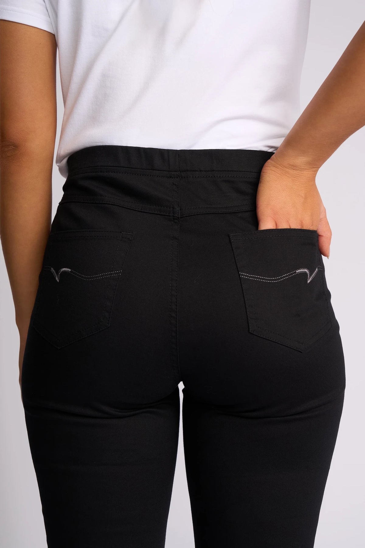 A person is wearing the Vassalli Bay Pull On - Black slim-leg jeans and a white shirt, with their right hand placed in the back pocket of the jeans. The image focuses on the lower back and hips.