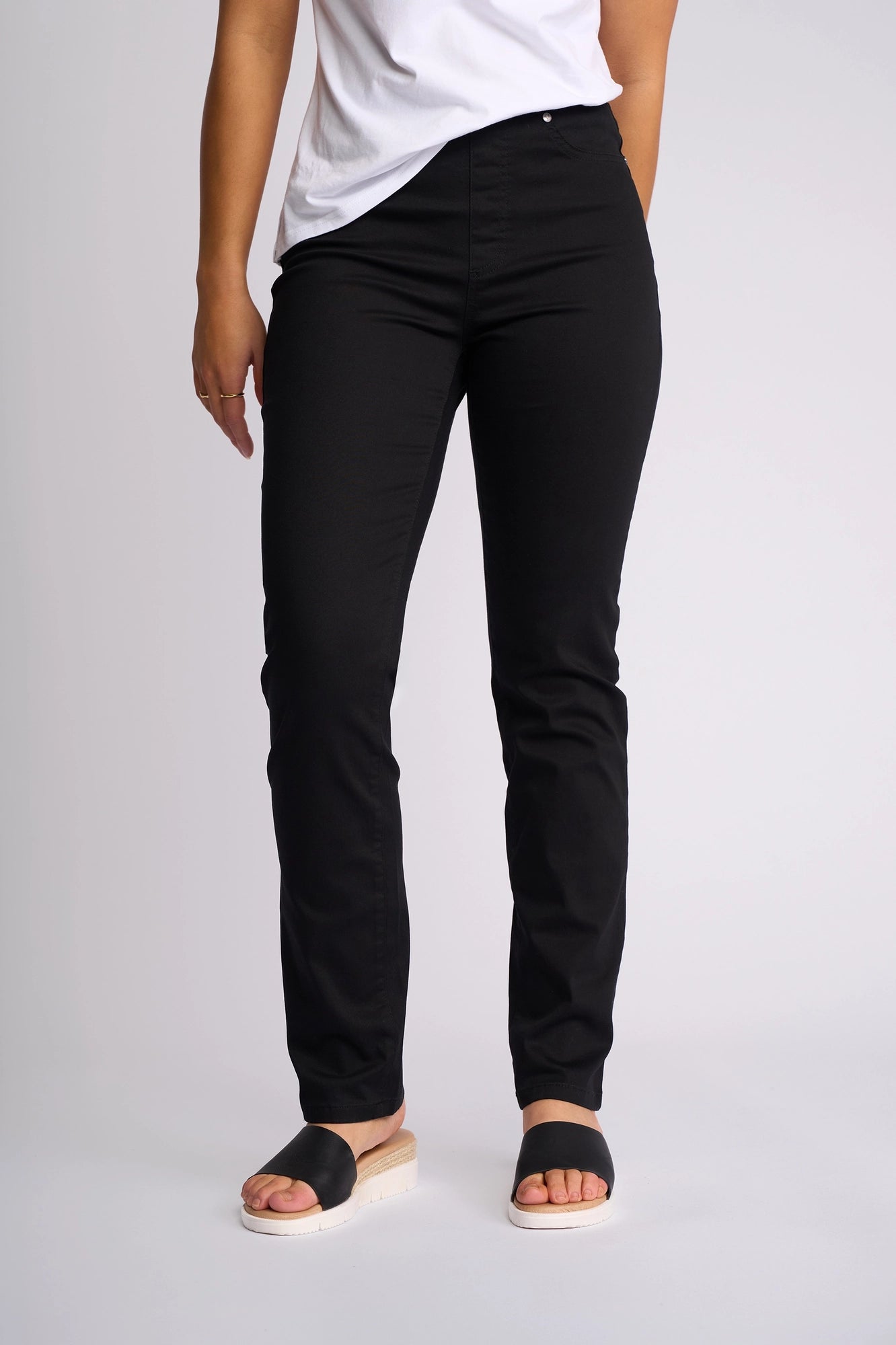 A person wearing a white shirt, Vassalli Bay Pull On pants in black, and black slide sandals stands against a plain light background. The image shows their body from the shoulders down.