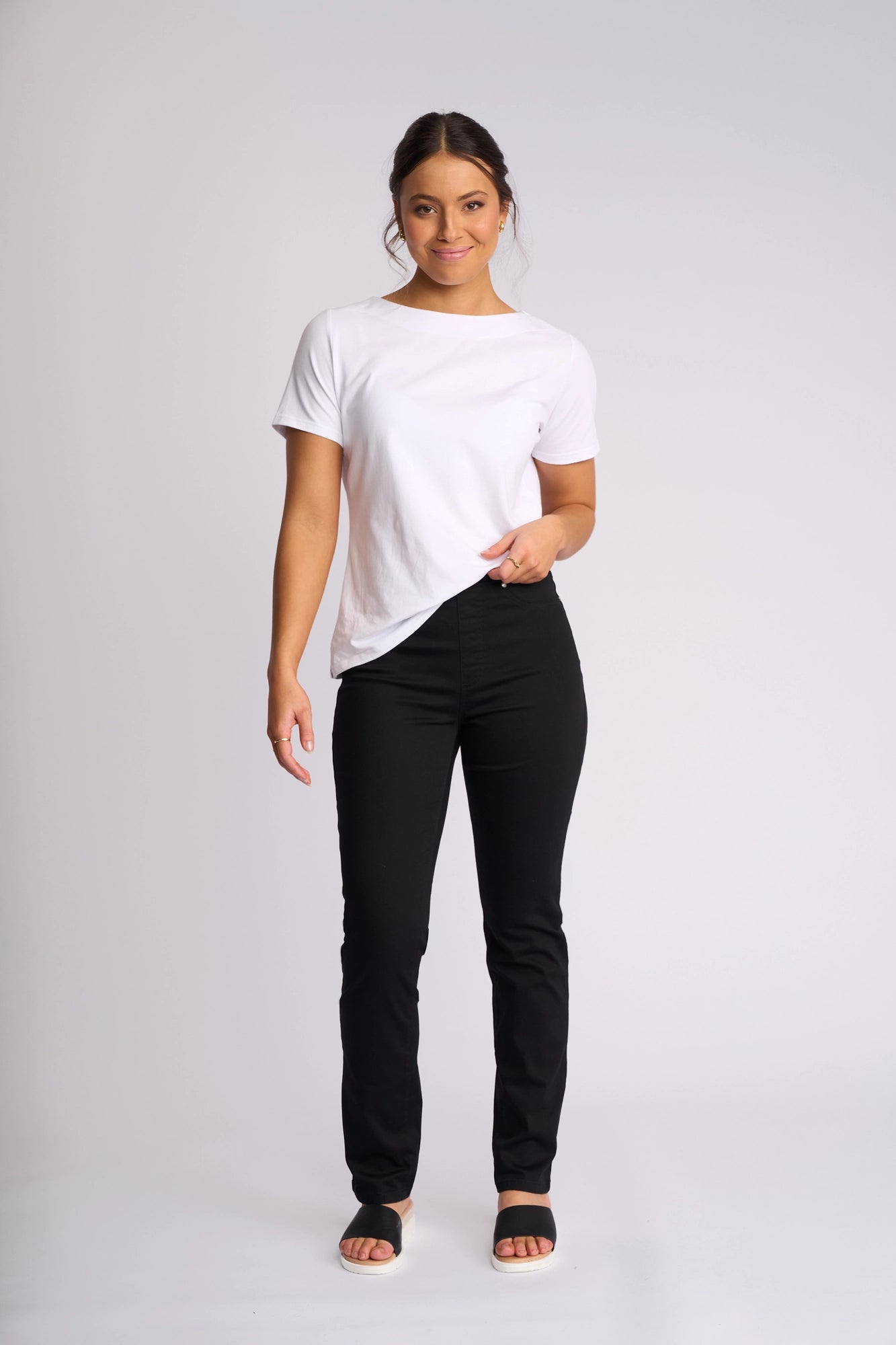 A woman is standing and smiling, wearing a white t-shirt and the Vassalli Bay Pull On - Black slim-leg pants in lightweight stretch fabric, paired with black sandals against a plain light background.