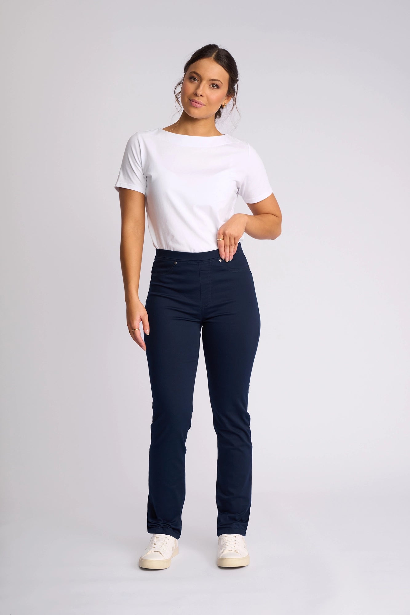 A woman stands against a plain background, wearing a white t-shirt, Vassalli’s Bay Pull On - Navy slim leg pant crafted from stretch fabric, and white sneakers. Her hair is tied back, and she has one hand slightly raised toward her waist.