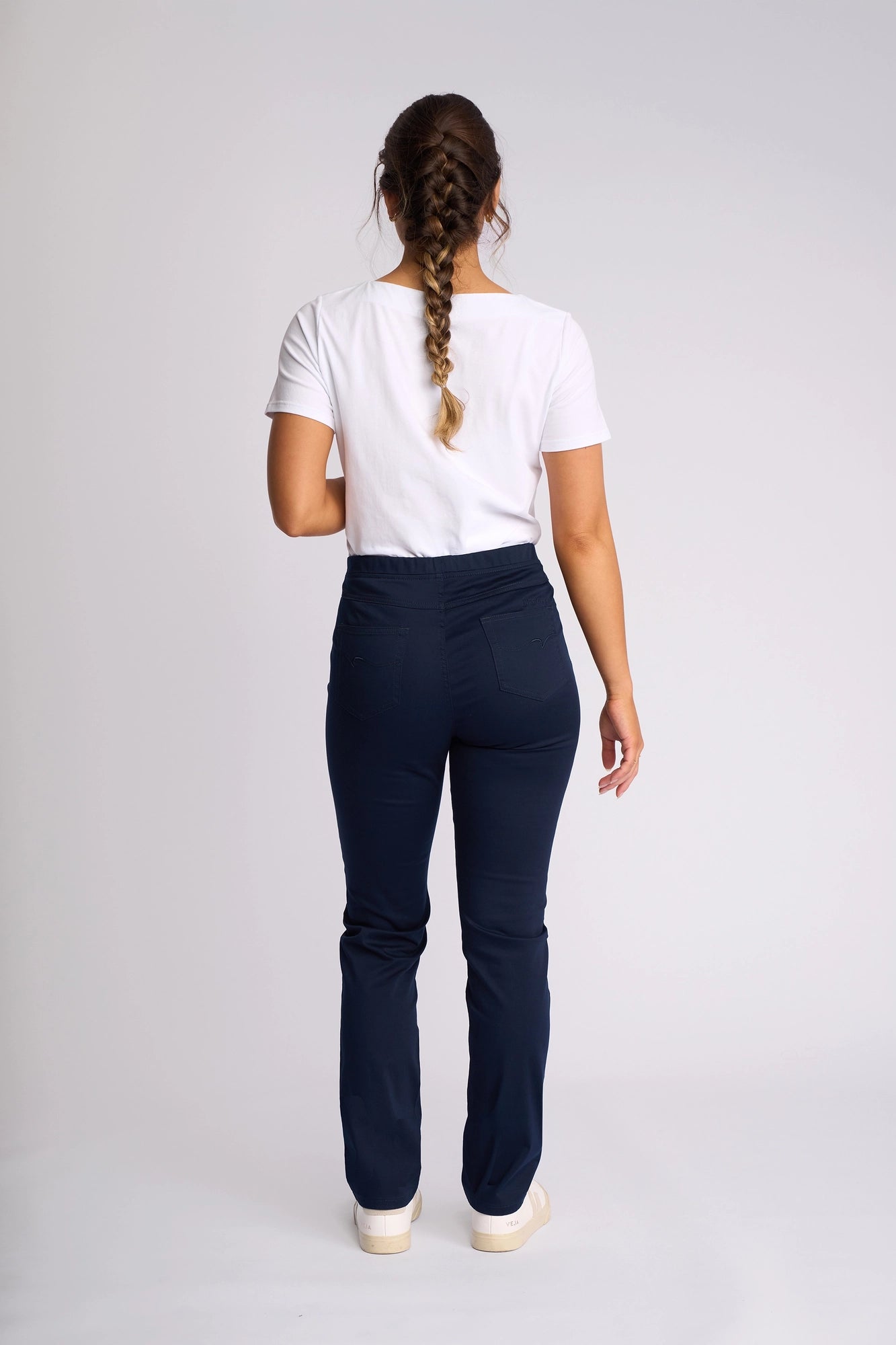 A woman with a long braid, wearing a white t-shirt and Vassalli’s Bay Pull On navy slim leg pants crafted from stretch fabric, stands facing away from the camera against a plain white background. She completes her look with crisp white shoes.
