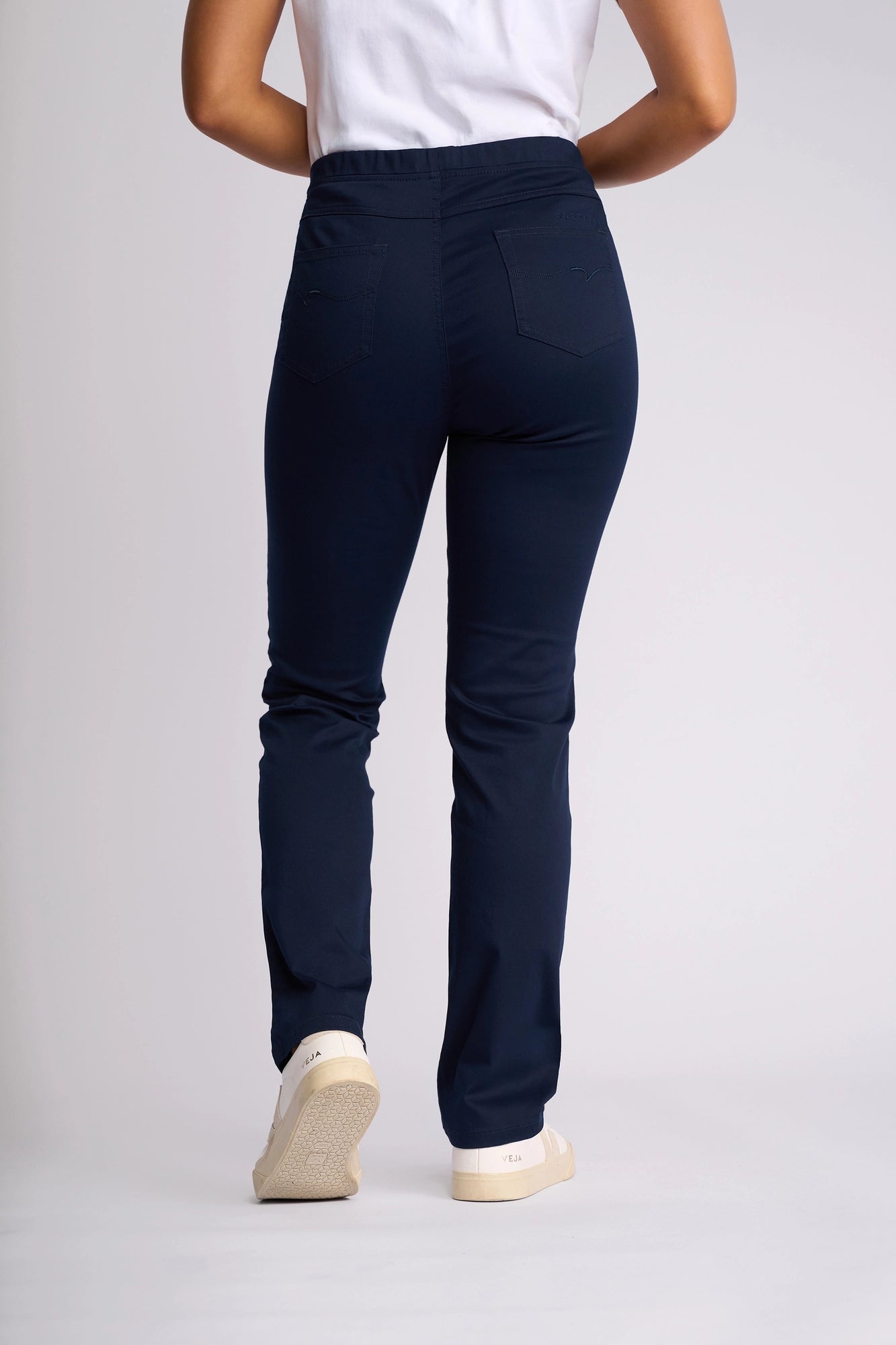 A person is standing with their back facing the camera against a plain light background, wearing a white shirt, white sneakers, and Vassalli Bay Pull On pants in navy.