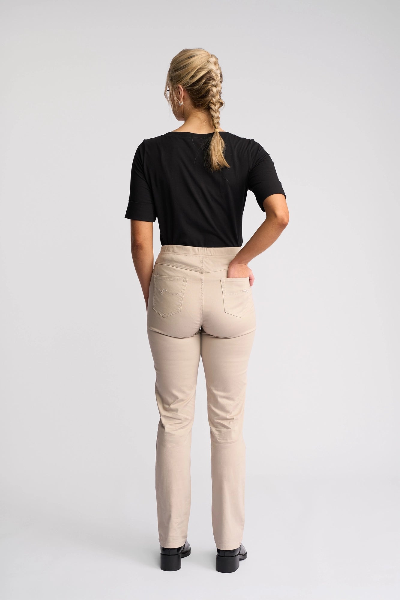 A woman with blond hair in a braid stands facing away, wearing a black short-sleeve shirt and Vassalli Bay Pull On - Pumice slim-leg pants crafted from stretch fabric, paired with black shoes against a plain light background.