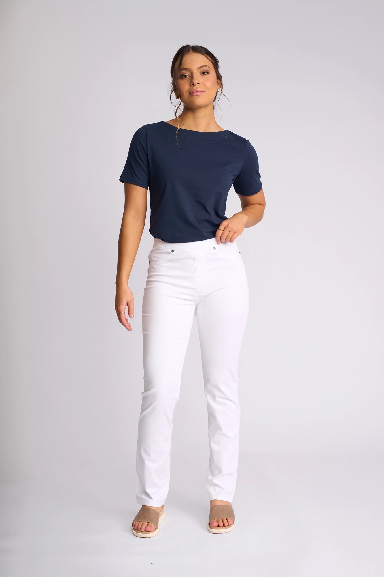 A woman stands against a plain light background wearing a navy blue short-sleeve top, beige sandals, and the Vassalli Bay Pull On - White slim-leg pants crafted from comfortable stretch fabric. Her hair is tied back and she gazes slightly to the side with a neutral expression.