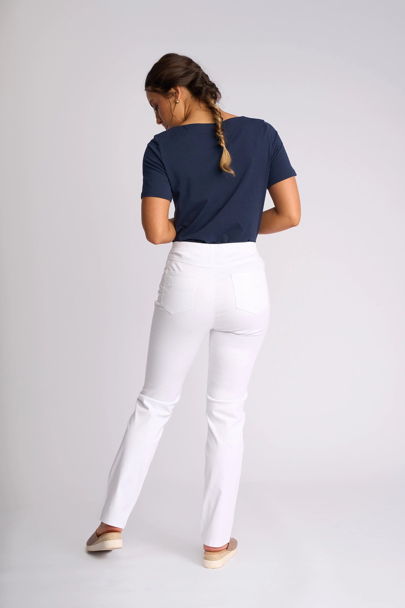 A woman with a braid wears a navy t-shirt, Vassalli Bay Pull On - White slim-leg pants, and slip-on shoes, standing with her back to the camera against a plain light background.