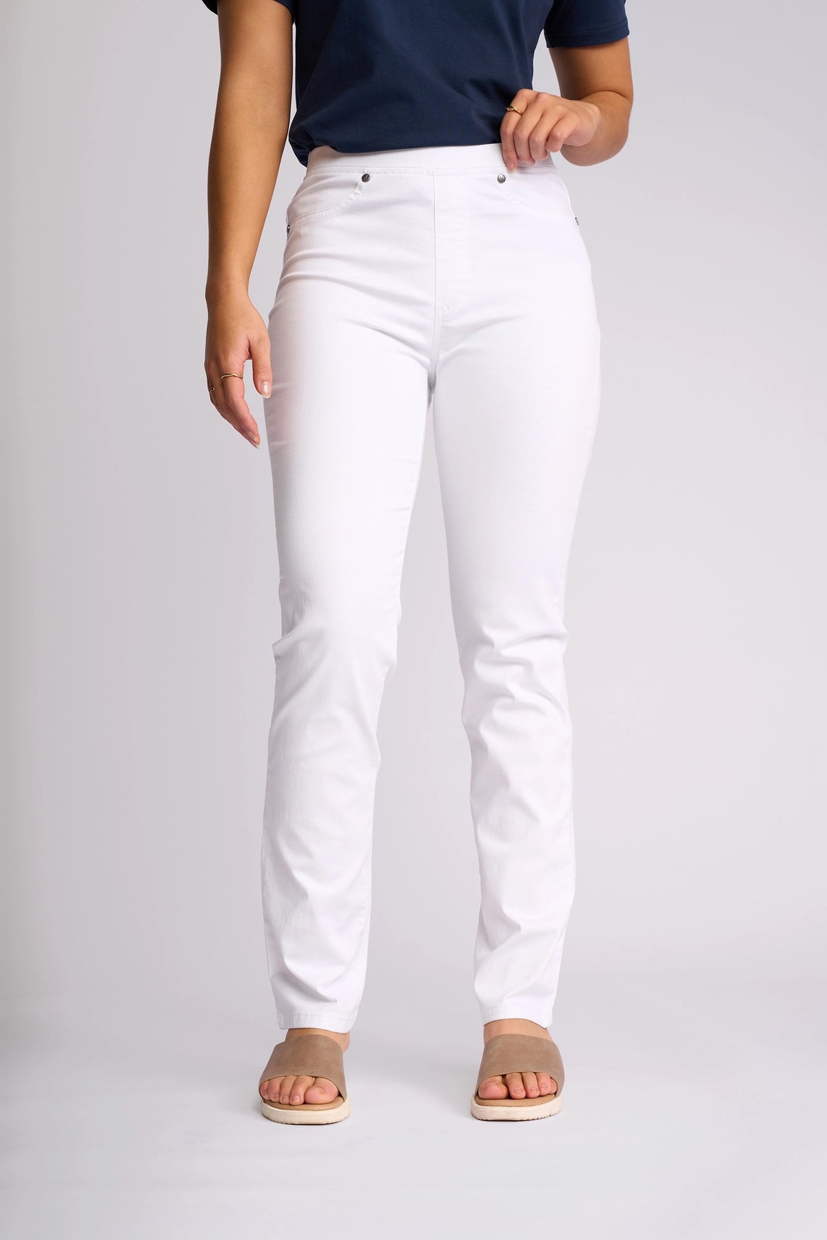 A person wearing a navy blue top, Vassalli’s Bay Pull On slim-leg pants in white, and beige sandals stands against a plain light background.