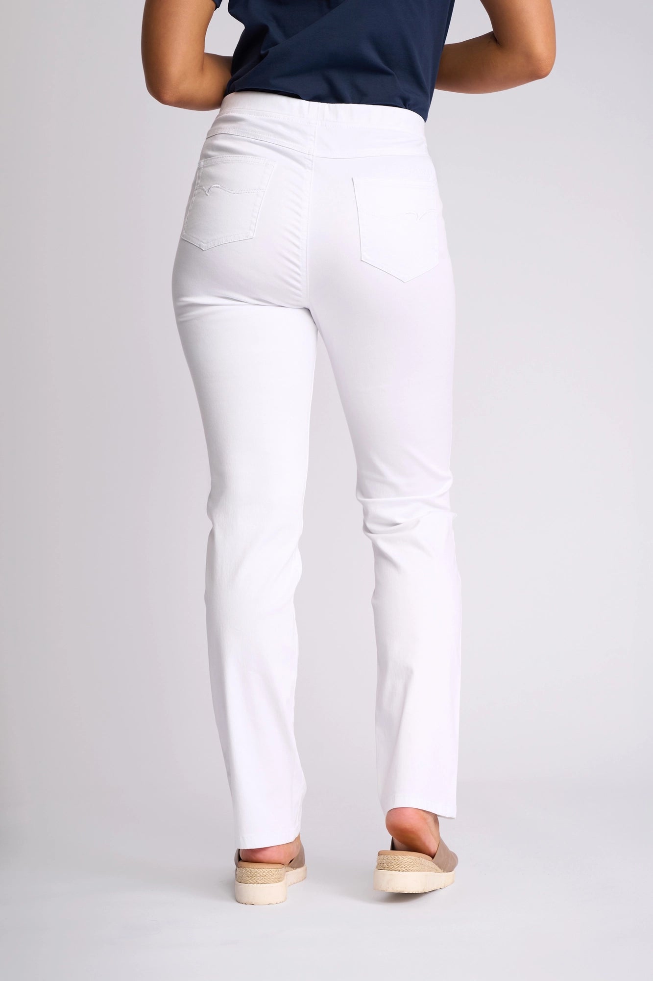 A person stands facing away against a plain light background, wearing Vassalli’s Bay Pull On pants in white with a slim-leg fit, paired with a navy shirt and beige shoes.