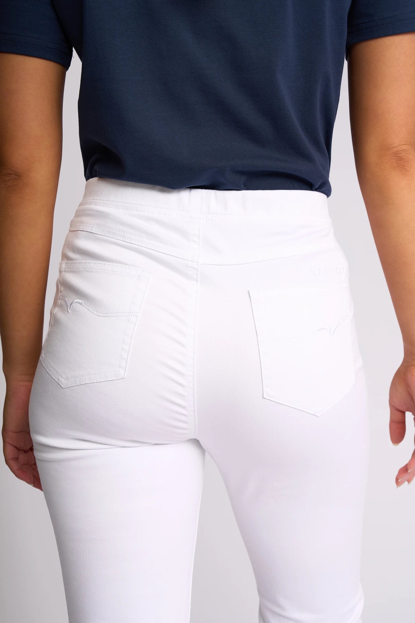 A person is shown from the back, focusing on the waist and hip area, wearing Vassalli’s Bay Pull On - White slim-leg pants. These pull-on style jeans feature two back pockets with stitched detailing and are crafted from comfortable stretch fabric for a flattering fit.