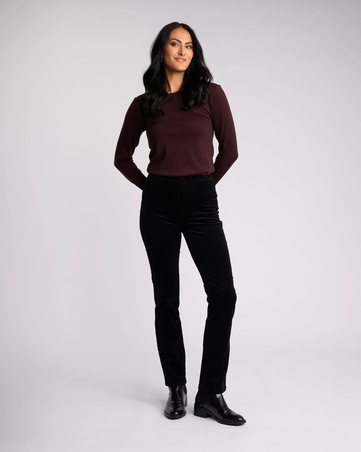 A woman with long dark hair, wearing the Vassalli Bay Cord Pull On pants in black and a maroon long-sleeve top, stands against a plain light background, smiling with her hands behind her back.