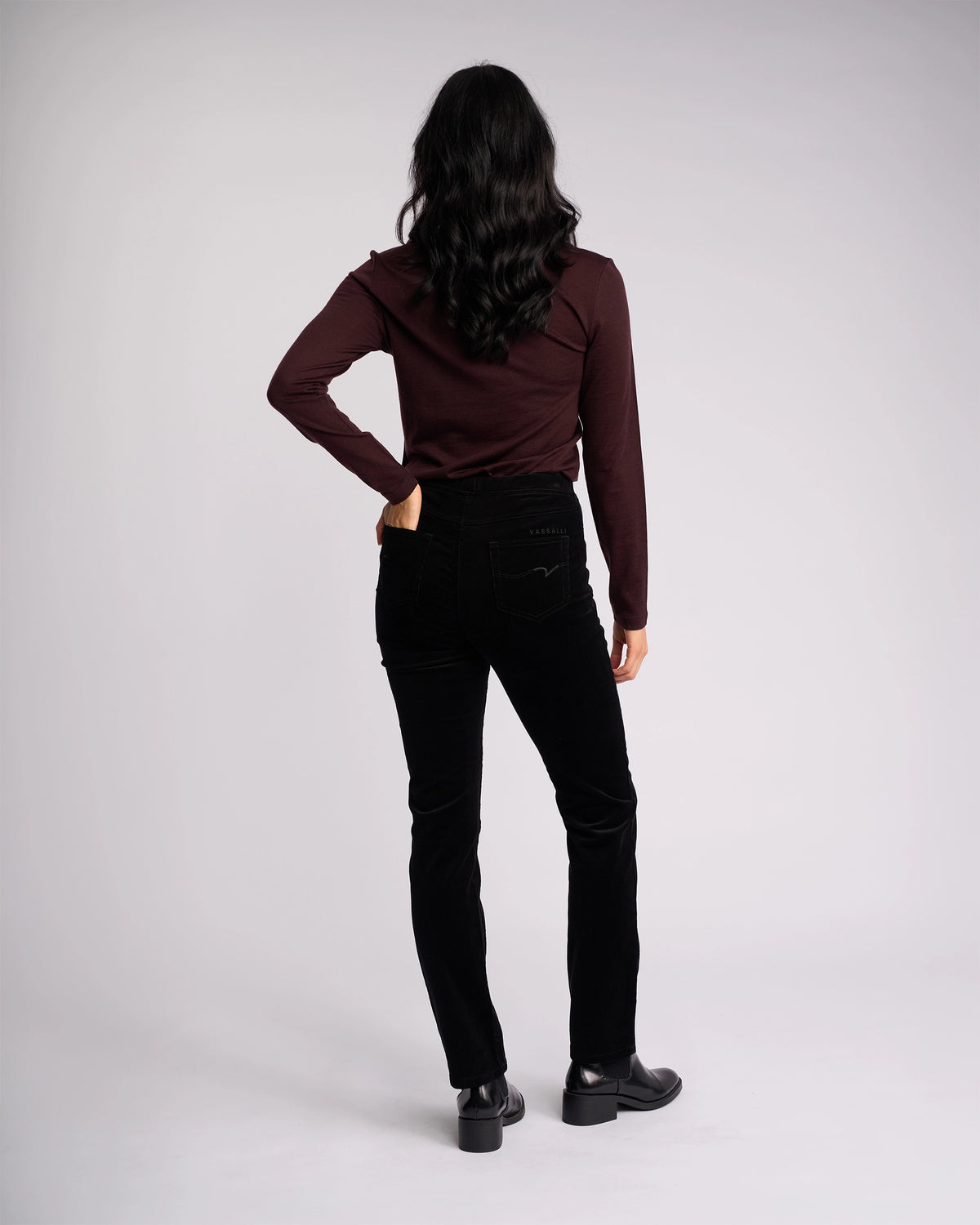 A person with long dark hair wears a maroon long-sleeve top and Vassalli Bay Cord Pull On - Black pants, standing with their back to the camera against a plain light background.