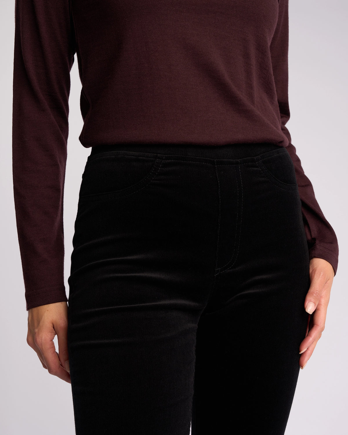 A person models the Vassalli Bay Cord Pull On - Black, paired with a long-sleeved dark maroon top. Only their torso and upper legs are visible against a plain light background.