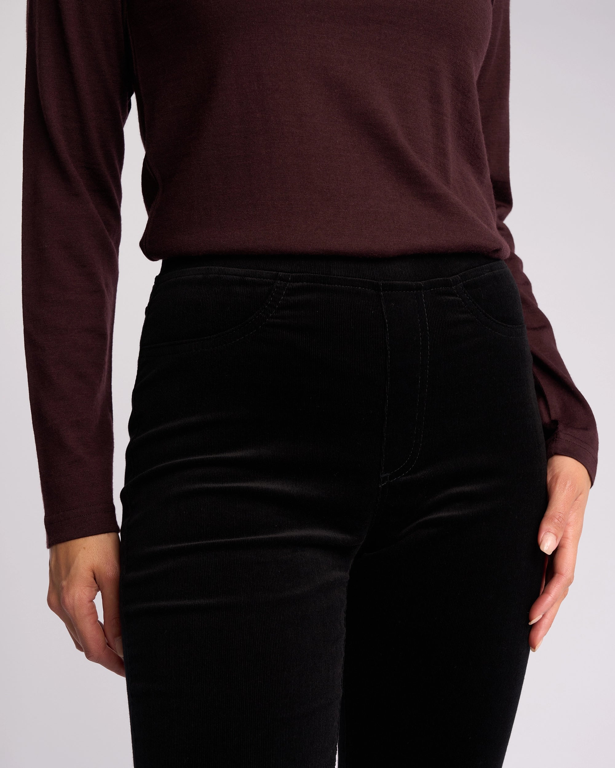 A person models the Vassalli Bay Cord Pull On - Black, paired with a long-sleeved dark maroon top. Only their torso and upper legs are visible against a plain light background.