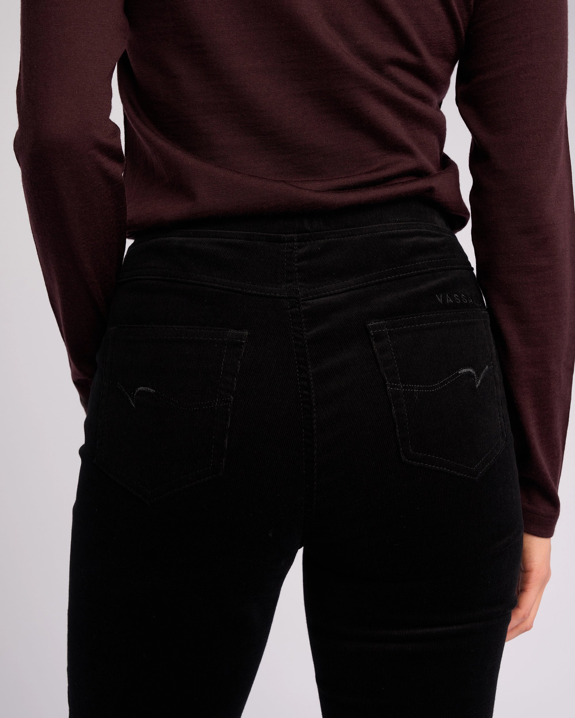 A person is shown from the back wearing Vassalli's Bay Cord Pull On pants in black, highlighting the high-waisted slim-leg fit at the waist and hips with both arms relaxed by their sides.