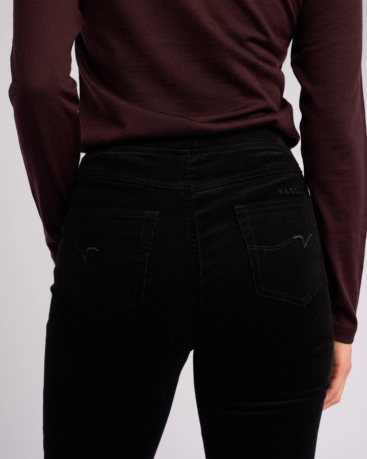 A person is shown from the back wearing Vassalli's Bay Cord Pull On pants in black, highlighting the high-waisted slim-leg fit at the waist and hips with both arms relaxed by their sides.