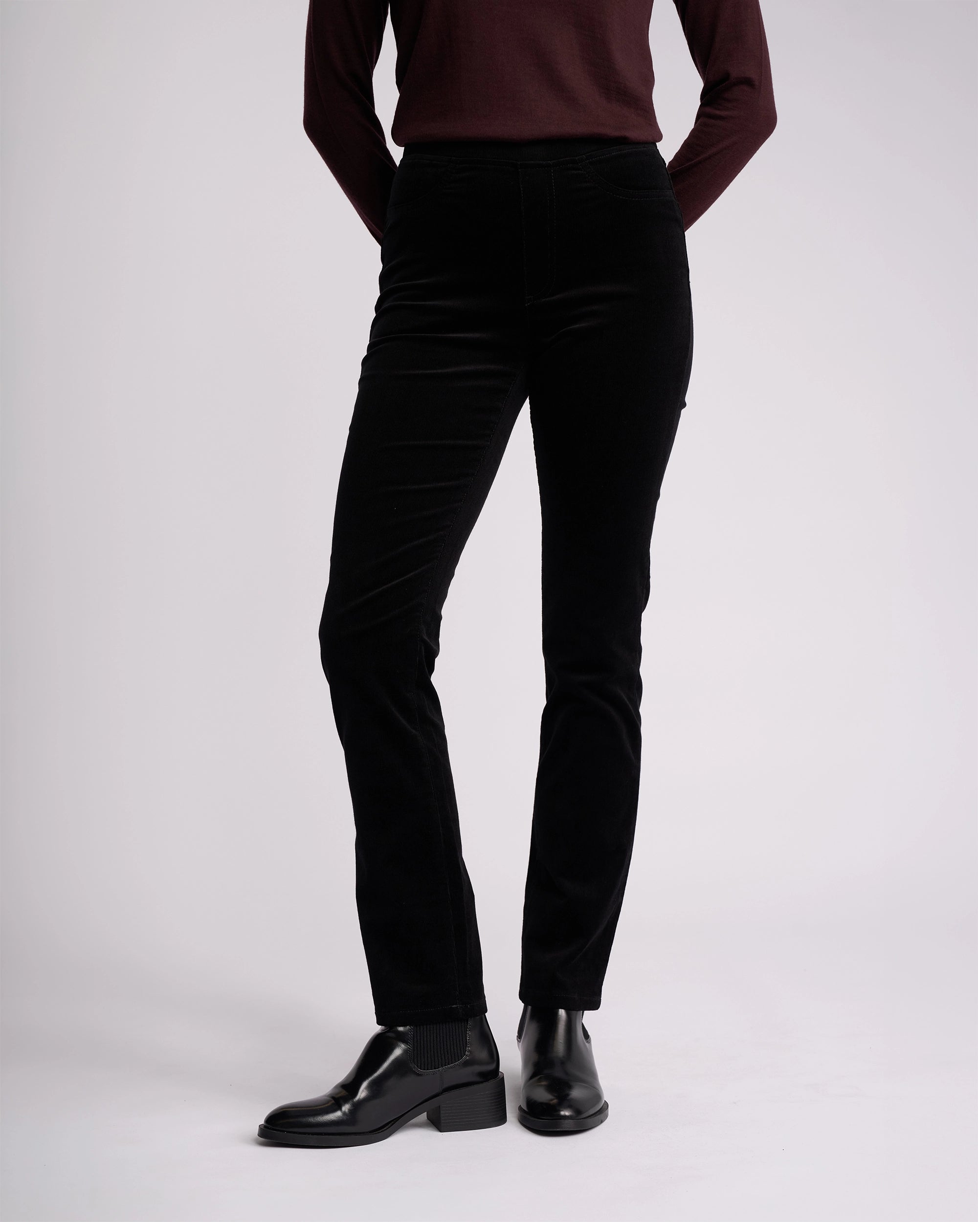 A person models the Vassalli Bay Cord Pull On - Black pants, paired with a dark burgundy long-sleeve top and black ankle boots, standing hands behind their back against a plain light background. Their face is not shown.
