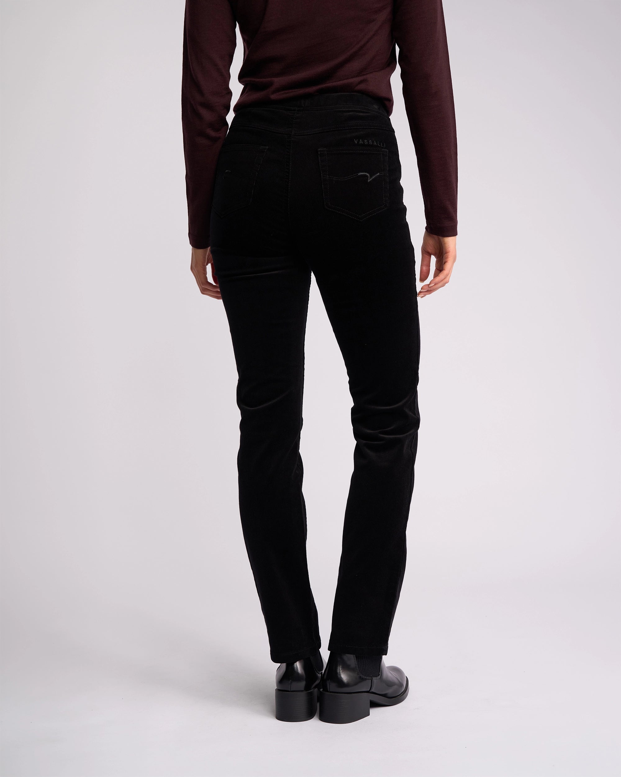 A person stands facing away in a long-sleeve dark burgundy top, Vassalli Bay Cord Pull On - Black pants, and black shoes against a plain light background.