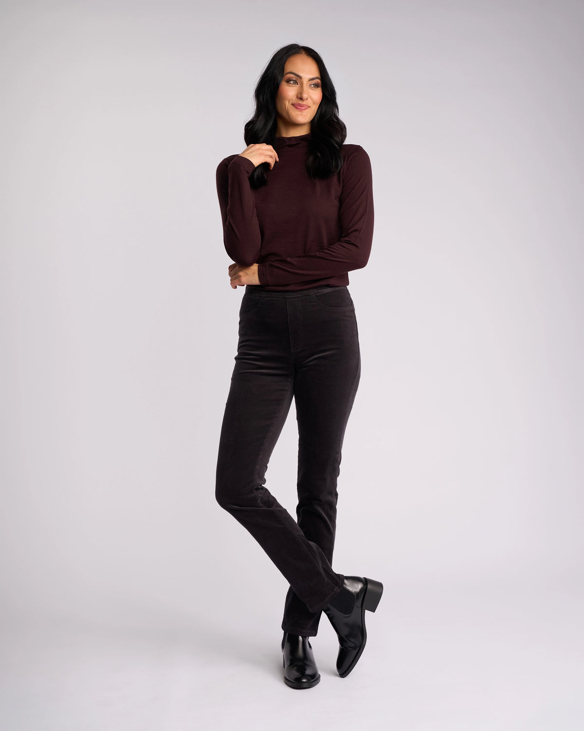 A woman with long dark hair smiles and poses against a plain light background, wearing Vassalli's Bay Cord Pull On pants in Charcoal paired with a maroon long-sleeve top and black ankle boots.
