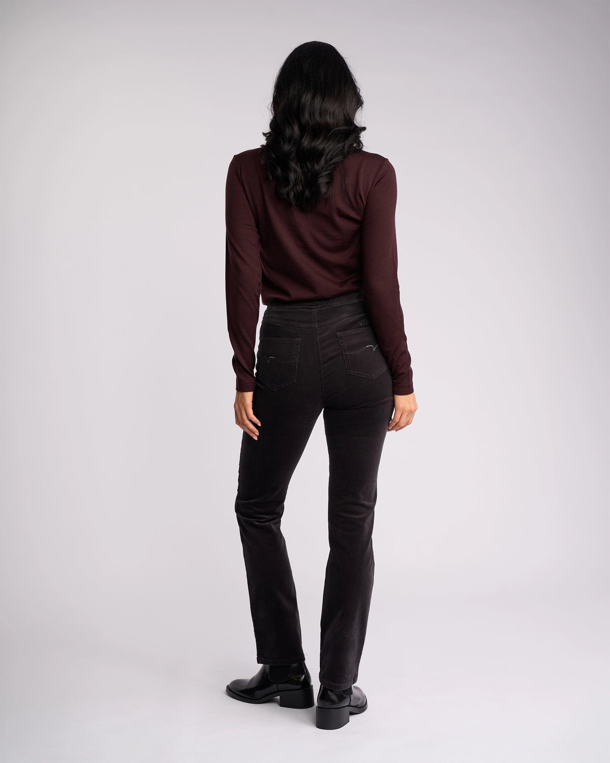A person with long, dark wavy hair stands facing away from the camera, wearing a long-sleeve burgundy top and Vassalli Bay Cord Pull On pants in Charcoal, paired with black shoes against a plain light gray background.