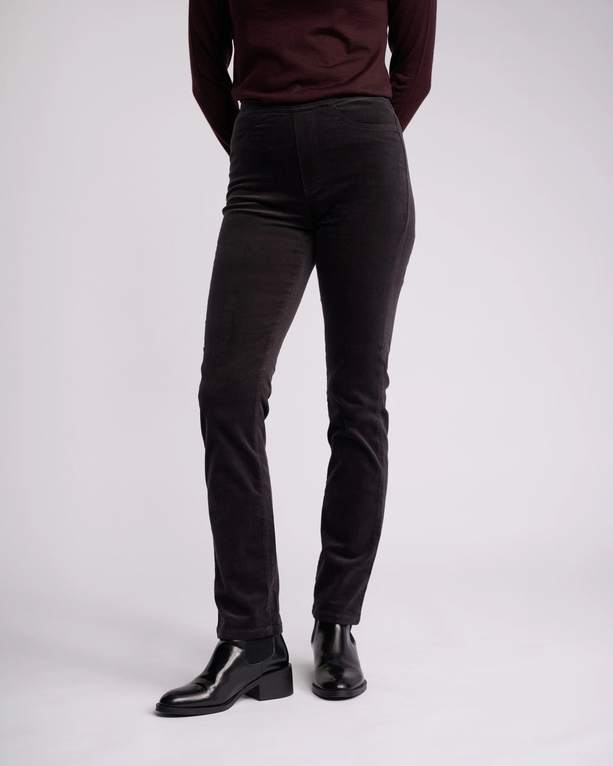 A person stands against a plain background wearing the Vassalli Bay Cord Pull On pants in charcoal with a dark maroon long-sleeve top and black ankle boots. Their arms are relaxed at their sides, and their head is out of the frame.