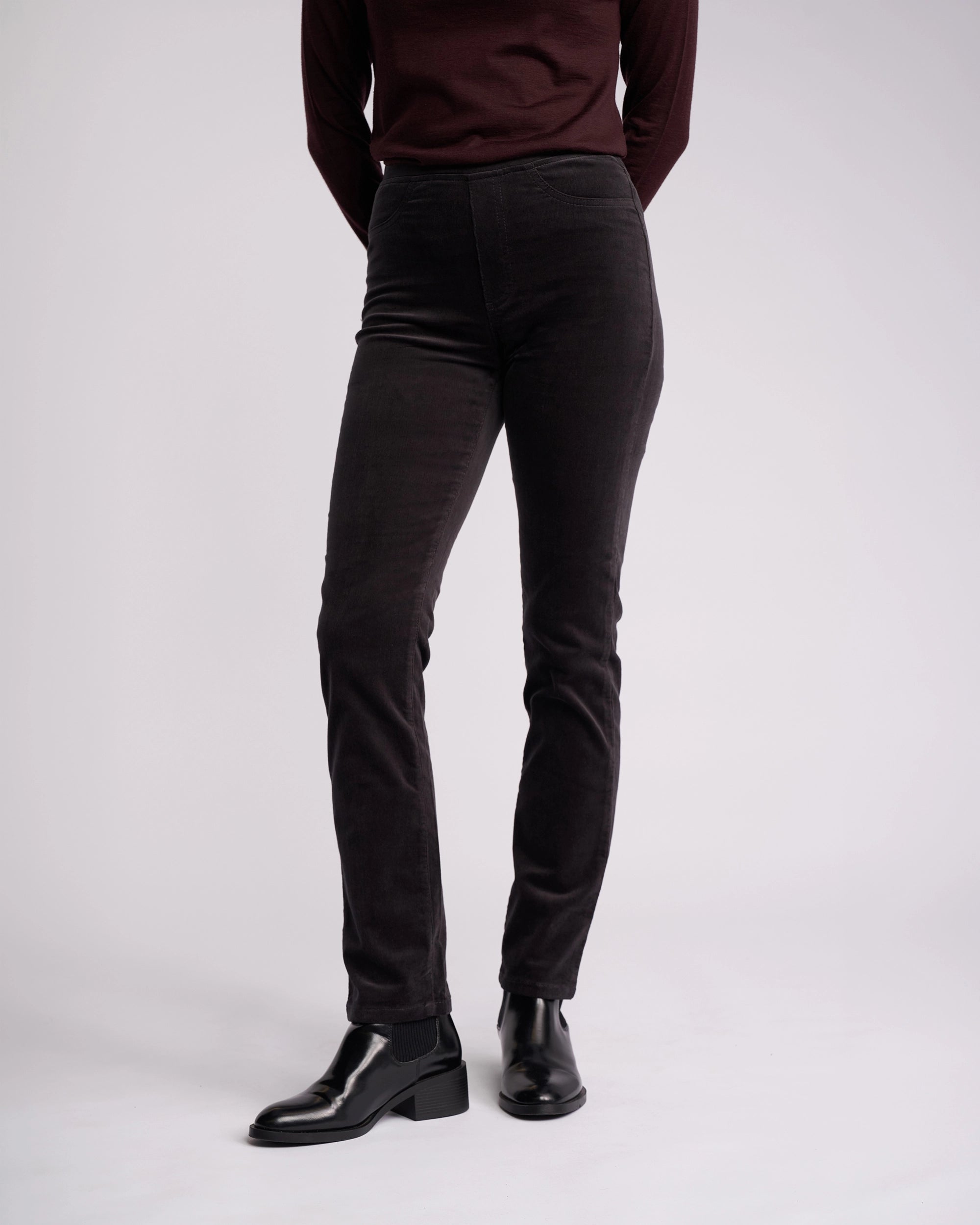A person stands against a plain background wearing the Vassalli Bay Cord Pull On pants in charcoal with a dark maroon long-sleeve top and black ankle boots. Their arms are relaxed at their sides, and their head is out of the frame.