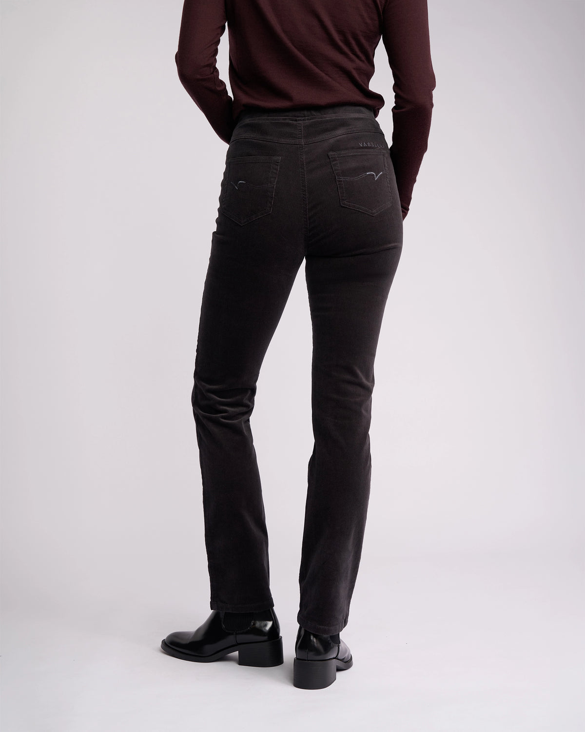 A person wearing a long-sleeve maroon top, Vassalli Bay Cord Pull On pants in charcoal, and black heeled boots stands with their back to the camera against a plain light background.