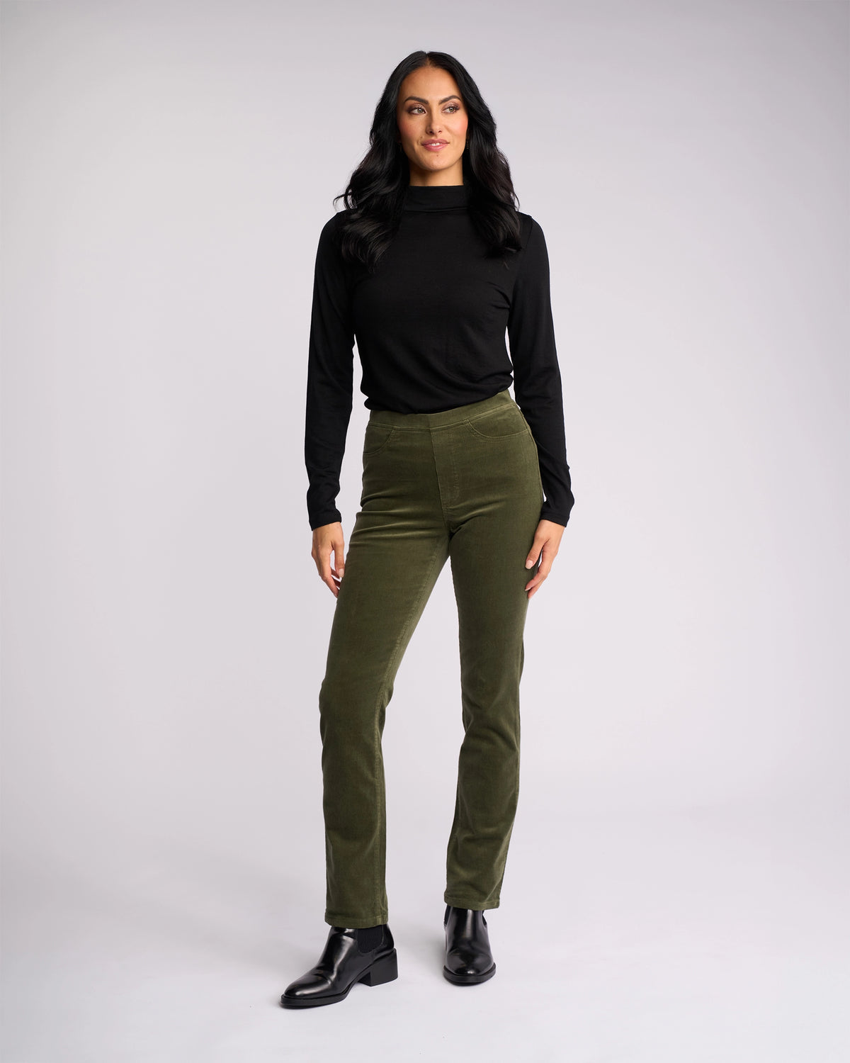 A woman with long dark hair stands confidently against a light background, wearing a black long-sleeve top, black ankle boots, and Vassalli Bay Cord Pull On pants in Deep Sage.