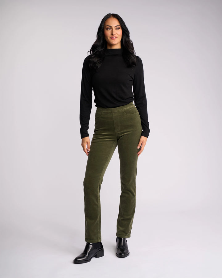 A woman with long dark hair stands confidently against a light background, wearing a black long-sleeve top, black ankle boots, and Vassalli Bay Cord Pull On pants in Deep Sage.