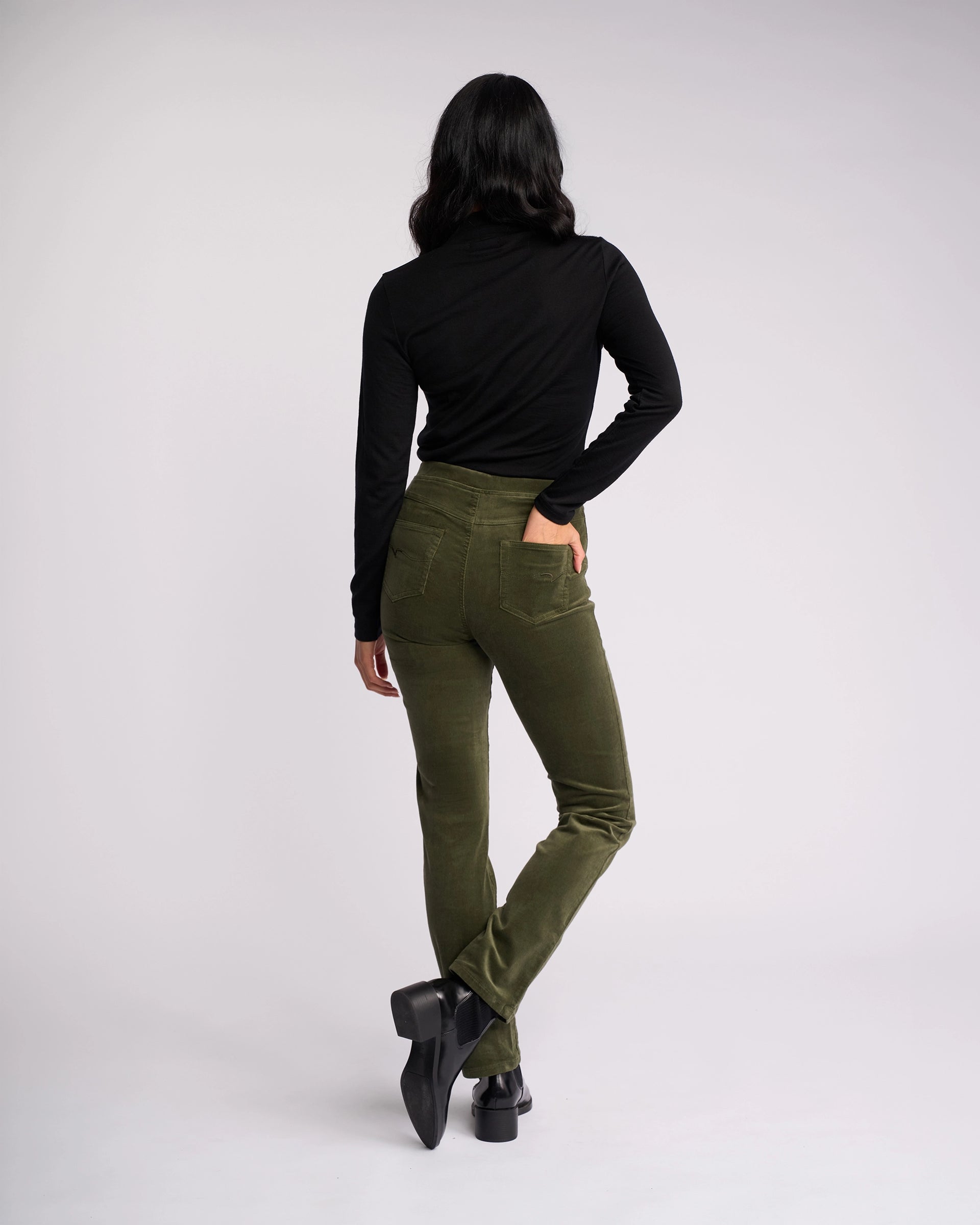 A person faces away from the camera, wearing a black long-sleeve top and Vassalli Bay Cord Pull On pants in Deep Sage, paired with black heeled shoes, one leg slightly crossed behind the other against a plain light background.