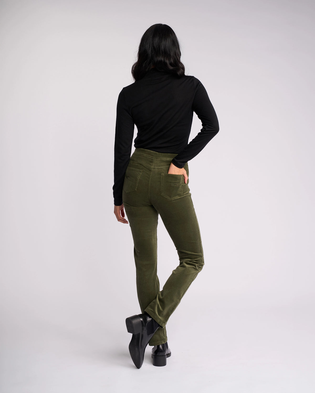 A person faces away from the camera, wearing a black long-sleeve top and Vassalli Bay Cord Pull On pants in Deep Sage, paired with black heeled shoes, one leg slightly crossed behind the other against a plain light background.