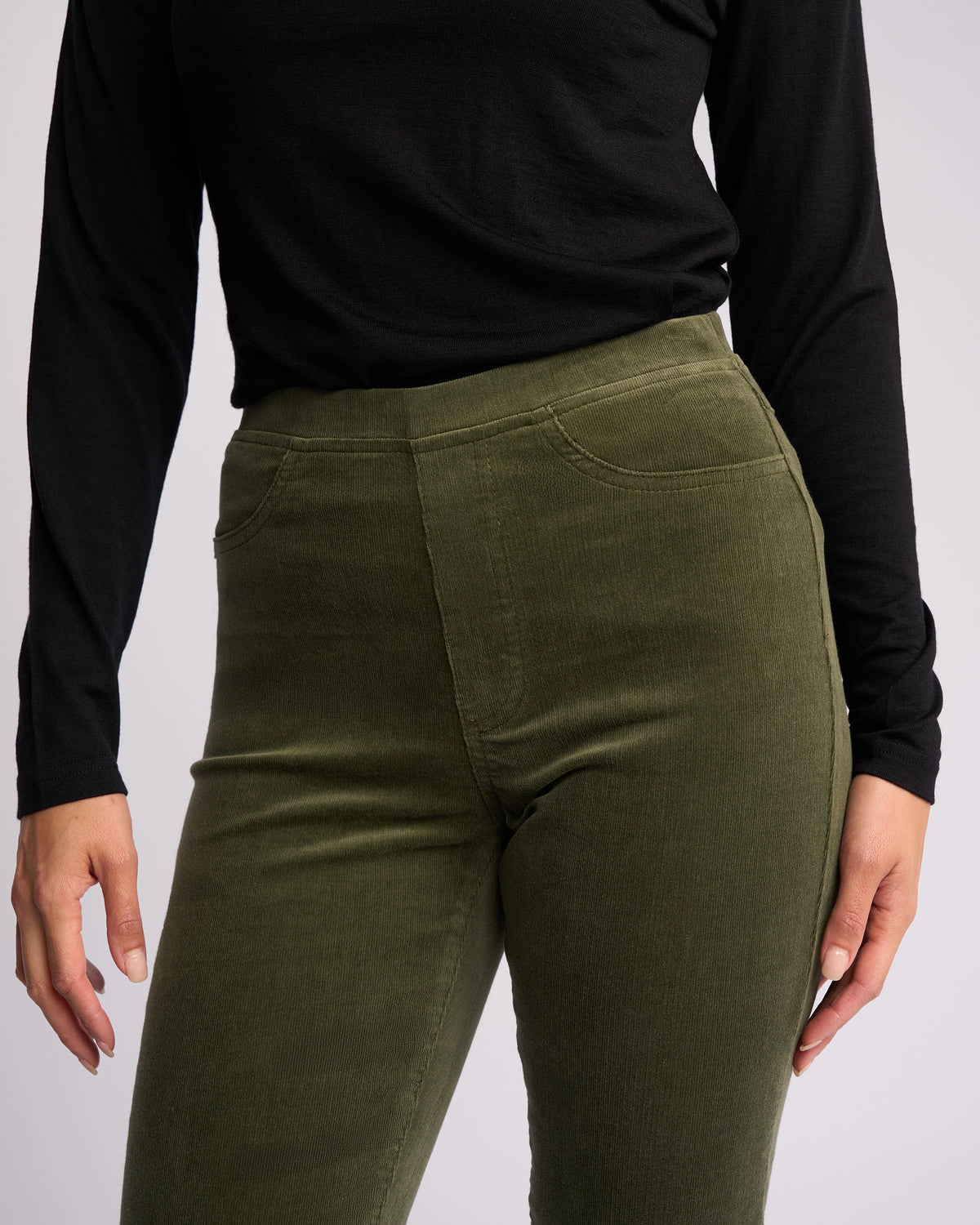A person wears Vassalli's Bay Cord Pull On in Deep Sage, featuring a high-waisted, slim-leg fit and subtle stretch pinwale cord for style and comfort, standing against a neutral background. Image cropped from shoulders to thighs.