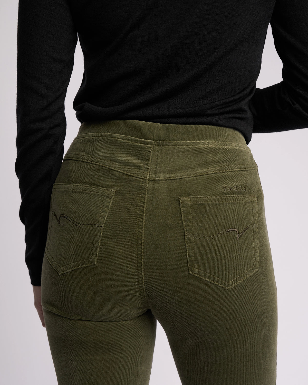 A person shown from the back wears Vassalli's Bay Cord Pull On pants in Deep Sage, paired with a black long-sleeve top, highlighting the waist and upper legs.