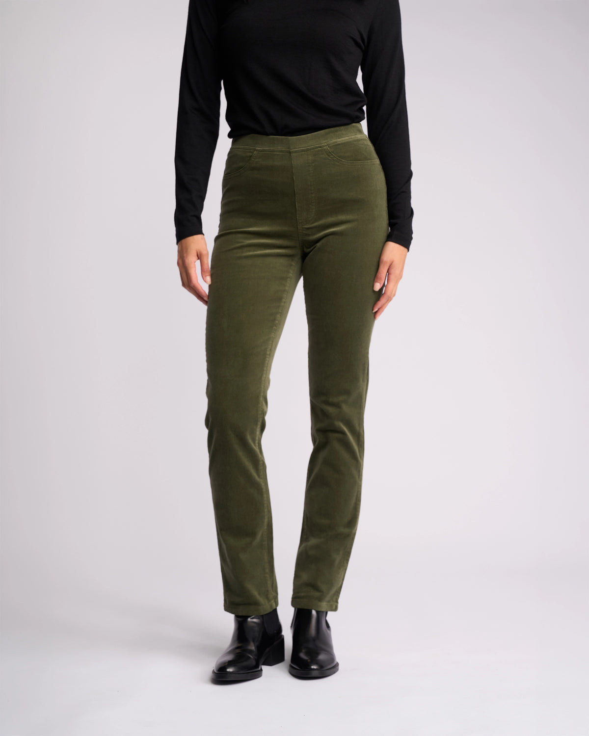 A person models the Vassalli Bay Cord Pull On - Deep Sage slim-leg pants, paired with an all-black long-sleeve top and black boots, standing against a plain white background.
