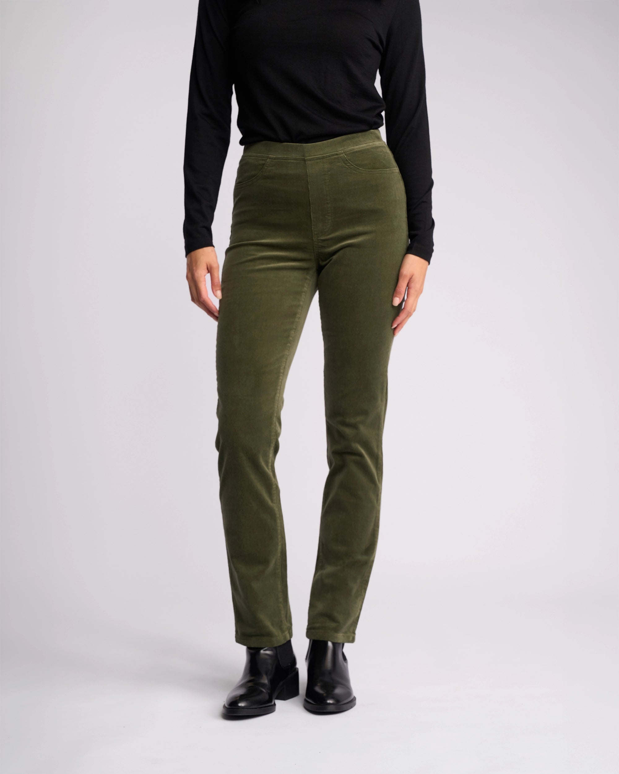 A person models the Vassalli Bay Cord Pull On - Deep Sage slim-leg pants, paired with an all-black long-sleeve top and black boots, standing against a plain white background.