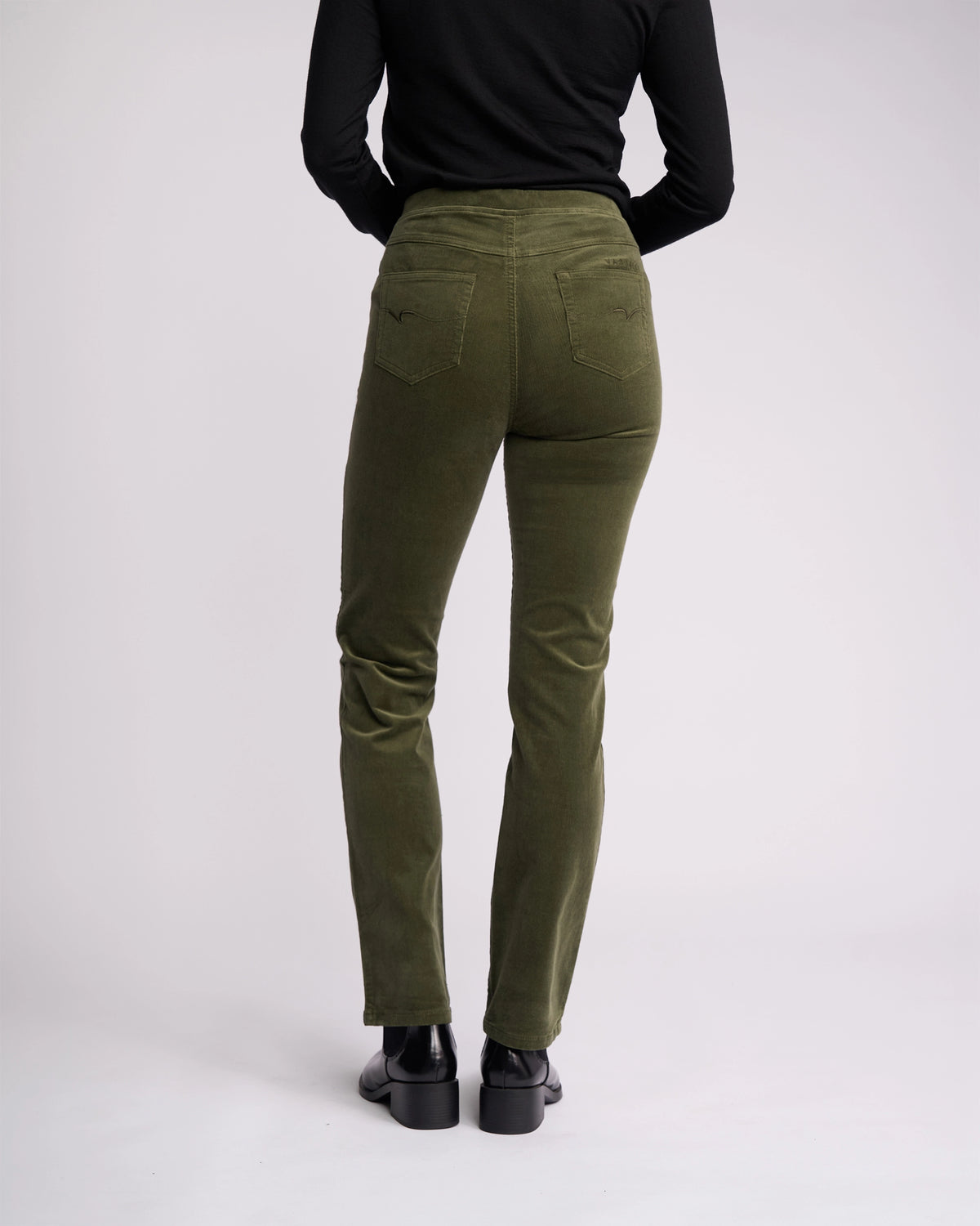 A person stands facing away from the camera against a plain light background wearing Vassalli Bay Cord Pull On pants in Deep Sage, paired with a black long-sleeve top and black shoes.