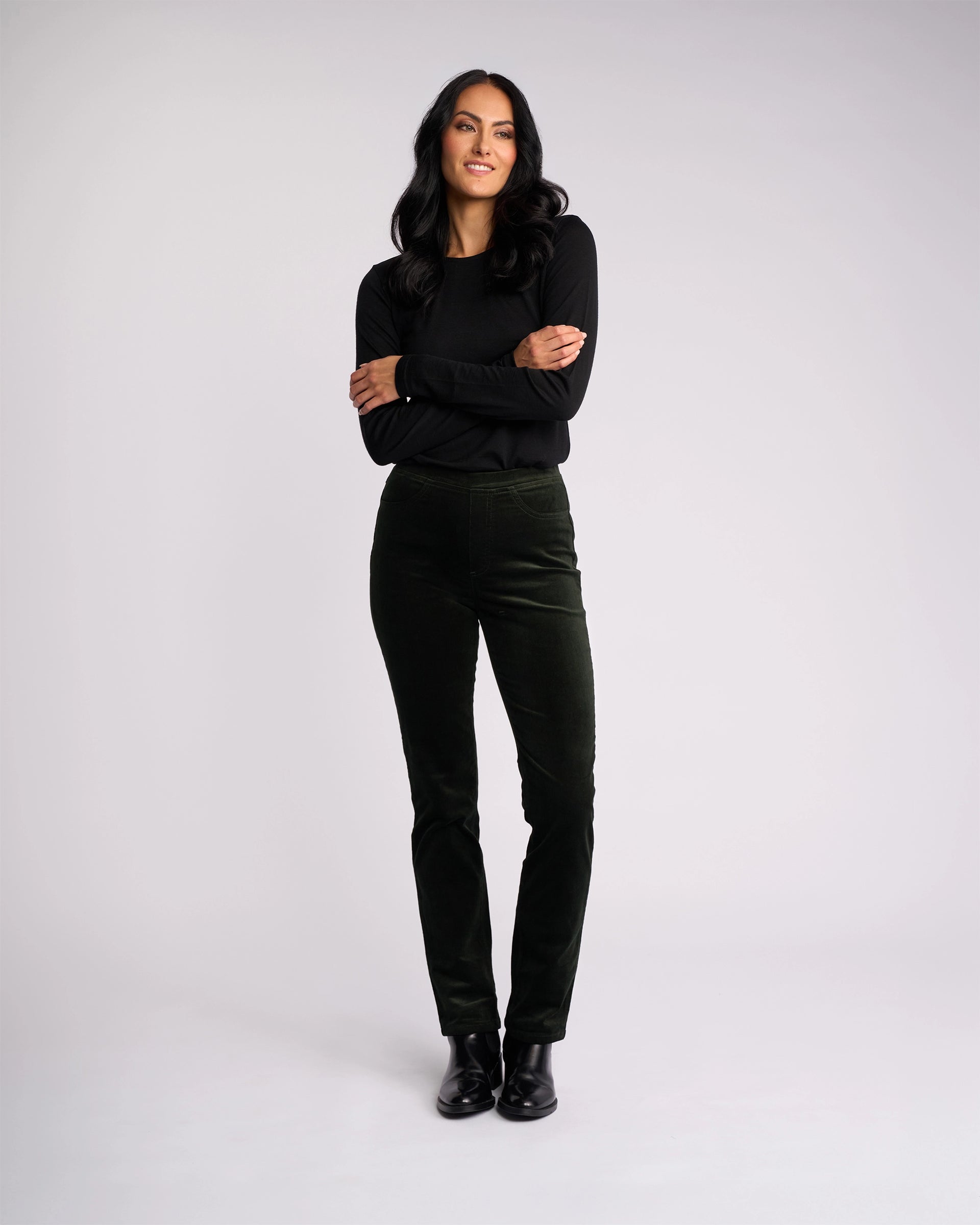 A woman with long dark hair stands against a light background, smiling with her arms crossed. She wears a black top, black shoes, and the Vassalli Bay Cord Pull On - Forest pants for a modern, polished look.