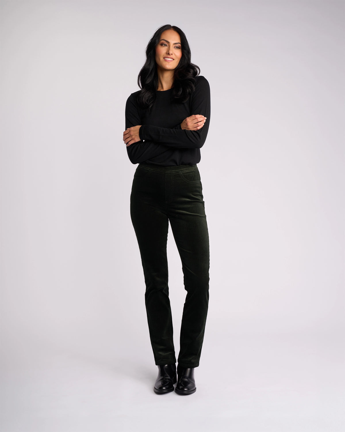 A woman with long dark hair stands against a light background, smiling with her arms crossed. She wears a black top, black shoes, and the Vassalli Bay Cord Pull On - Forest pants for a modern, polished look.