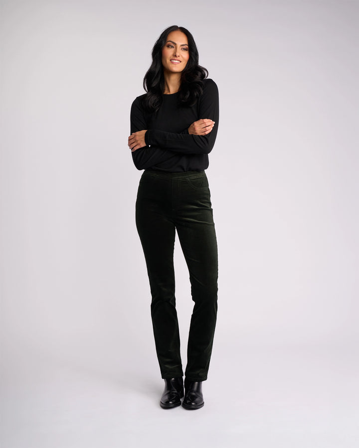 A woman with long dark hair stands against a light background, smiling with her arms crossed. She wears a black top, black shoes, and the Vassalli Bay Cord Pull On - Forest pants for a modern, polished look.