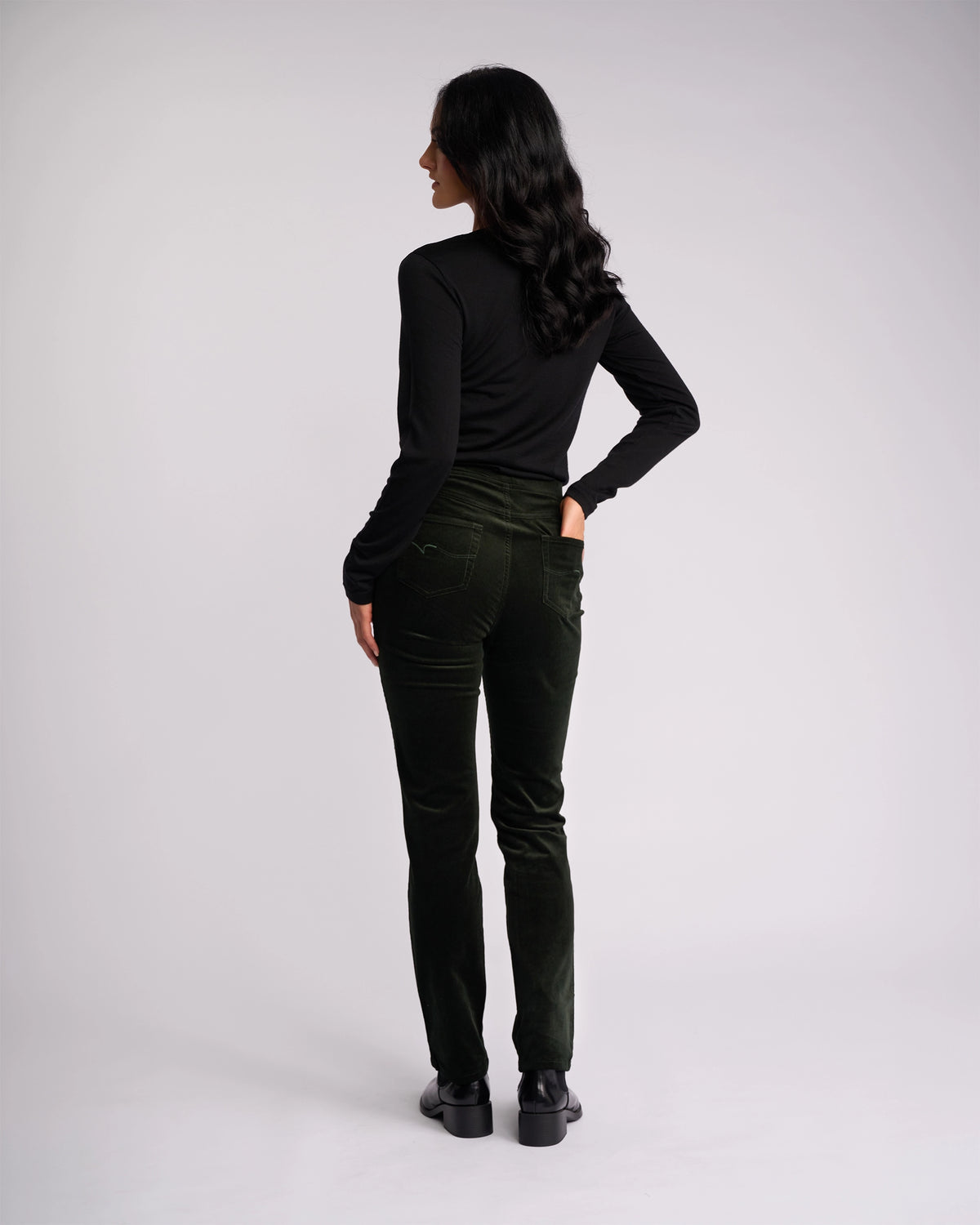 A woman with long dark hair stands facing away, wearing a black long-sleeve top, black shoes, and Vassalli Bay Cord Pull On pants in Forest against a plain light background.