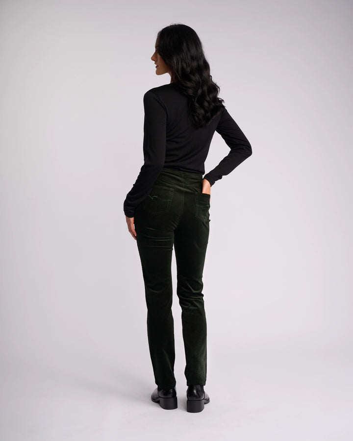 A woman with long dark hair stands facing away, wearing a black long-sleeve top, black shoes, and Vassalli Bay Cord Pull On pants in Forest against a plain light background.