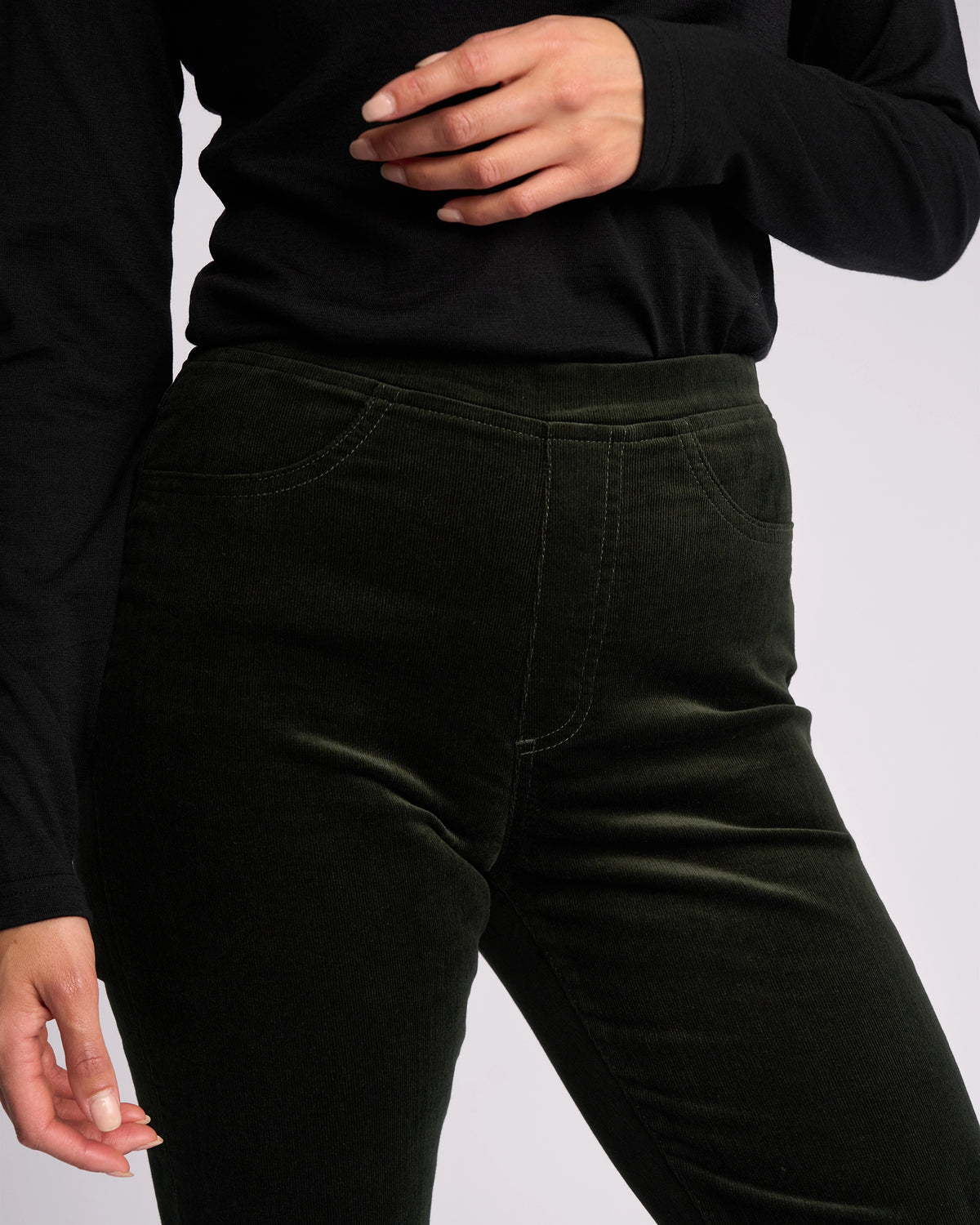 A person models Vassalli's Bay Cord Pull On pants in Forest, shown from shoulder to thigh, paired with a long-sleeve black top; one hand is on their hip and the other hangs by their side against a plain background.