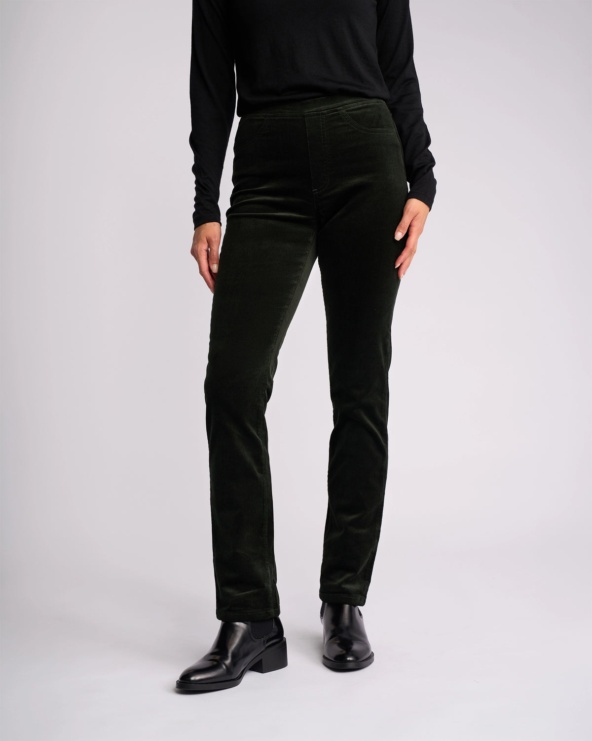 A person wears a black long-sleeve top and Vassalli Bay Cord Pull On - Forest pants, paired with black ankle boots, standing against a plain light background. Only the body from shoulders down is visible.