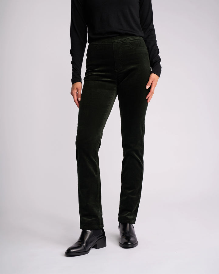 A person wears a black long-sleeve top and Vassalli Bay Cord Pull On - Forest pants, paired with black ankle boots, standing against a plain light background. Only the body from shoulders down is visible.