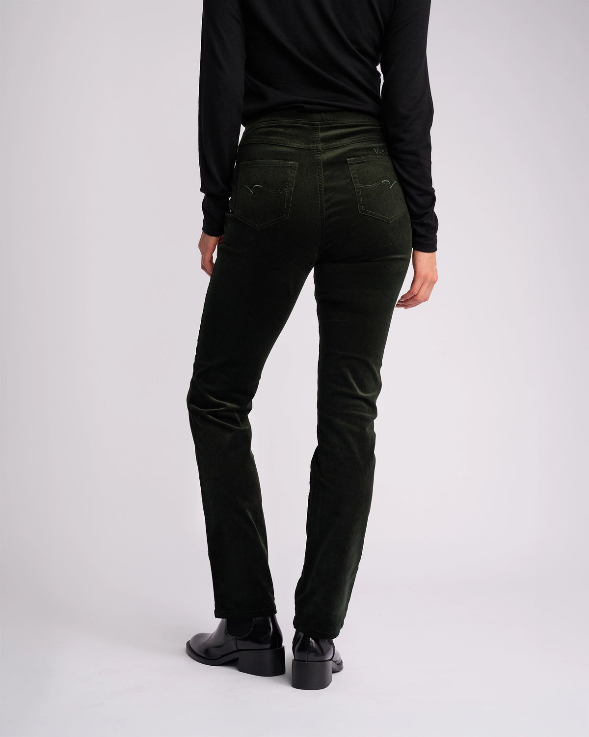 A person stands facing away against a plain light background, wearing Vassalli Bay Cord Pull On pants in Forest, a black long-sleeve top, and black shoes.