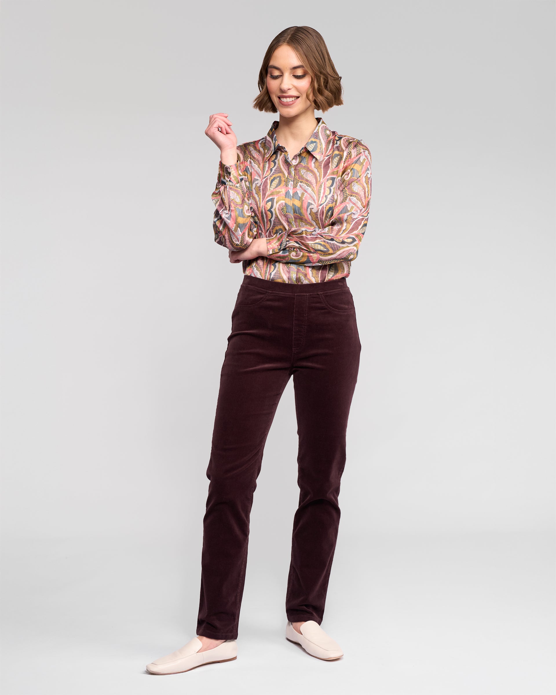 A woman stands against a plain light background, smiling and looking to the side with one arm raised. She wears maroon Vassalli Bay Cord Pull On - Merlot pants, a patterned long-sleeve blouse, and light-colored flats.