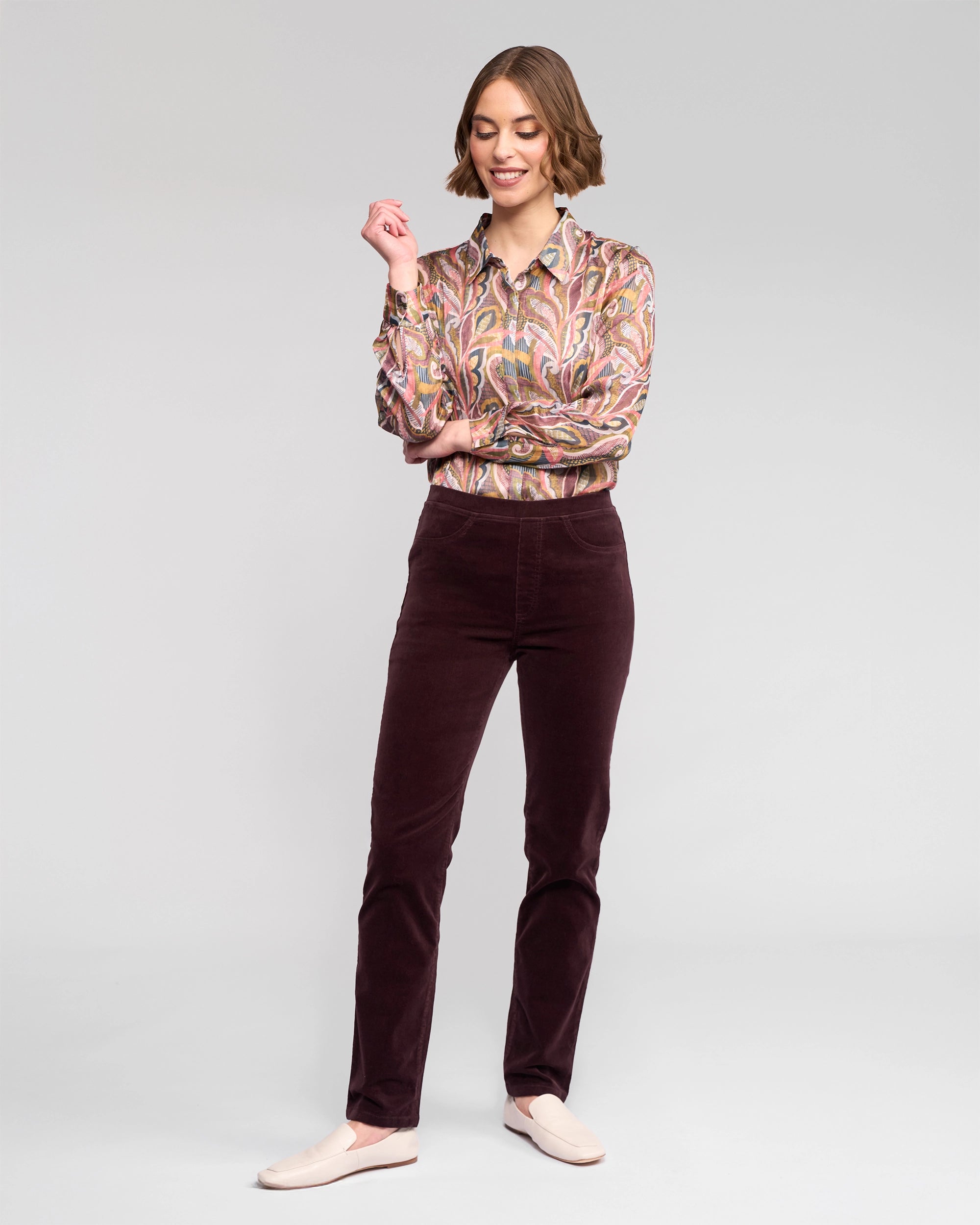 A woman stands confidently against a light gray background, smiling with one hand slightly raised, wearing Vassalli Bay Cord Pull On pants in Merlot.